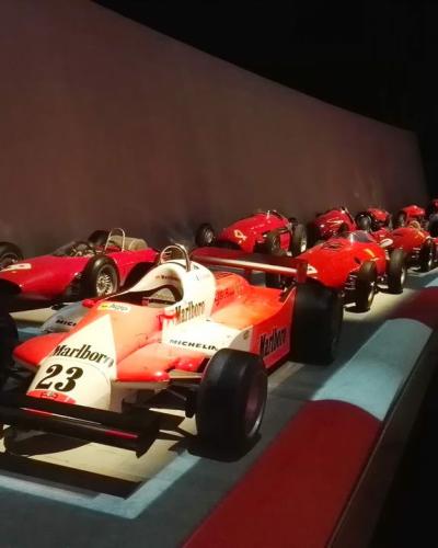 A row of red Formula 1 race cars (including a prominent Marlboro-liveried car with number 23) displayed on a track-themed floor inside a dimly lit museum gallery.