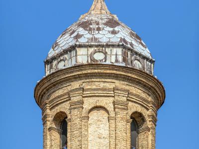 A close-up view of the top section of a brick bell tower (campanile) with a scalloped tile dome and a cross finial against a clear blue sky.