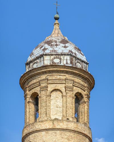 A close-up view of the top section of a brick bell tower (campanile) with a scalloped tile dome and a cross finial against a clear blue sky.