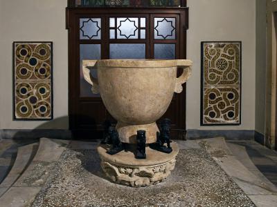 A large ancient stone basin (likely a baptismal font) supported by lion statues, situated indoors against a wall with decorative mosaic panels and a wooden door with geometric glass.