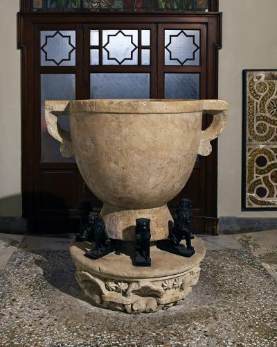 A large ancient stone basin (likely a baptismal font) supported by lion statues, situated indoors against a wall with decorative mosaic panels and a wooden door with geometric glass.