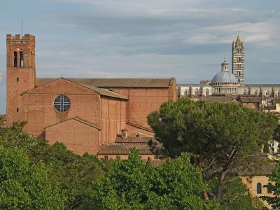 A view of Siena, Italy, featuring the red-brick Basilica of San Domenico in the foreground with its distinctive bell tower, and the Duomo di Siena (Siena Cathedral) with its striped facade and dome visible in the background.