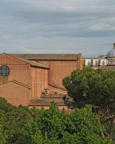 A view of Siena, Italy, featuring the red-brick Basilica of San Domenico in the foreground with its distinctive bell tower, and the Duomo di Siena (Siena Cathedral) with its striped facade and dome visible in the background.