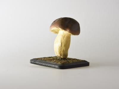 A realistic model of a Boletus edulis mushroom mounted on a small, dark, textured base against a plain white background.