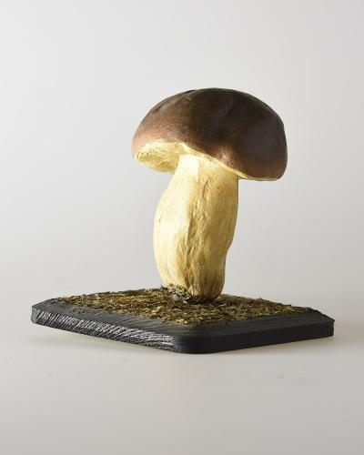 A realistic model of a Boletus edulis mushroom mounted on a small, dark, textured base against a plain white background.