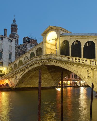 A stone arch bridge with a covered walkway featuring arched windows and shops, illuminated at dusk over a canal with wooden mooring posts in the foreground and historic buildings along the banks.