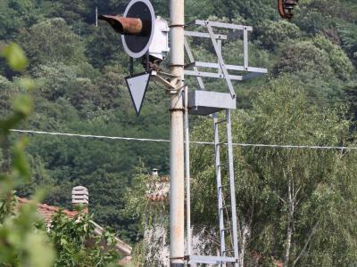 A close-up of a railway signal pole with a horn, triangular sign, and ladder structure, set against a backdrop of dense green trees and a glimpse of a building with a tiled roof.