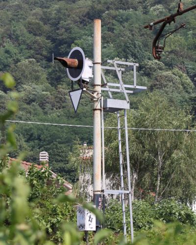 A close-up of a railway signal pole with a horn, triangular sign, and ladder structure, set against a backdrop of dense green trees and a glimpse of a building with a tiled roof.