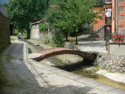 A small, low brick arch bridge spanning a shallow, rocky stream in a rural Tuscan setting, with stone walls, greenery, and a residential building with red flowers on the right.
