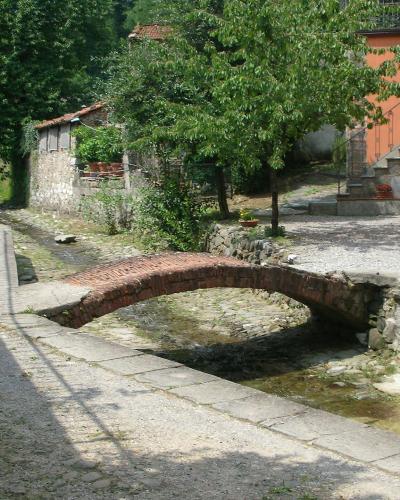A small, low brick arch bridge spanning a shallow, rocky stream in a rural Tuscan setting, with stone walls, greenery, and a residential building with red flowers on the right.