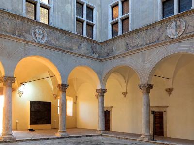 The image shows the interior courtyard (cortile) of a Renaissance palace, featuring a two-story loggia with stone columns, rounded arches, and windows on the upper level, with a black cat walking on the ground floor.