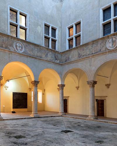 The image shows the interior courtyard (cortile) of a Renaissance palace, featuring a two-story loggia with stone columns, rounded arches, and windows on the upper level, with a black cat walking on the ground floor.