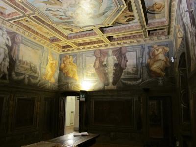 An interior room with elaborate Renaissance-style frescoes on the walls and a coffered ceiling, featuring allegorical figures, draped curtains, and architectural illusions; a wooden table sits in the foreground, and an exit sign is visible near a
