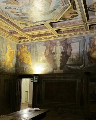An interior room with elaborate Renaissance-style frescoes on the walls and a coffered ceiling, featuring allegorical figures, draped curtains, and architectural illusions; a wooden table sits in the foreground, and an exit sign is visible near a