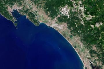 A satellite/aerial view of a coastal region showing a long sandy beach, urban development along the shore, green hills inland, and a prominent white quarry or exposed rock face on the hillside; no visible signage, people, or beach-level details.