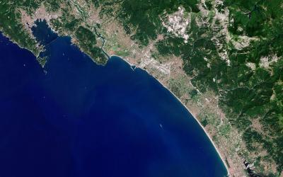 A satellite/aerial view of a coastal region showing a long sandy beach, urban development along the shore, green hills inland, and a prominent white quarry or exposed rock face on the hillside; no visible signage, people, or beach-level details.