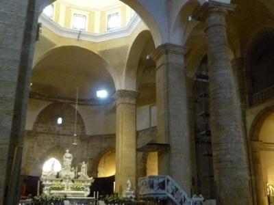 The interior of a large stone church with arched columns, wooden pews, an ornate altar with statues, and a white staircase leading to a pulpit or gallery.