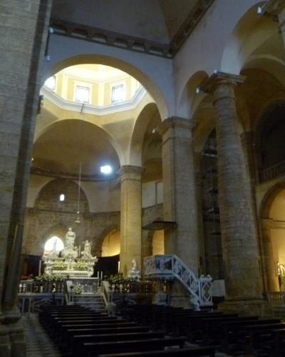 The interior of a large stone church with arched columns, wooden pews, an ornate altar with statues, and a white staircase leading to a pulpit or gallery.