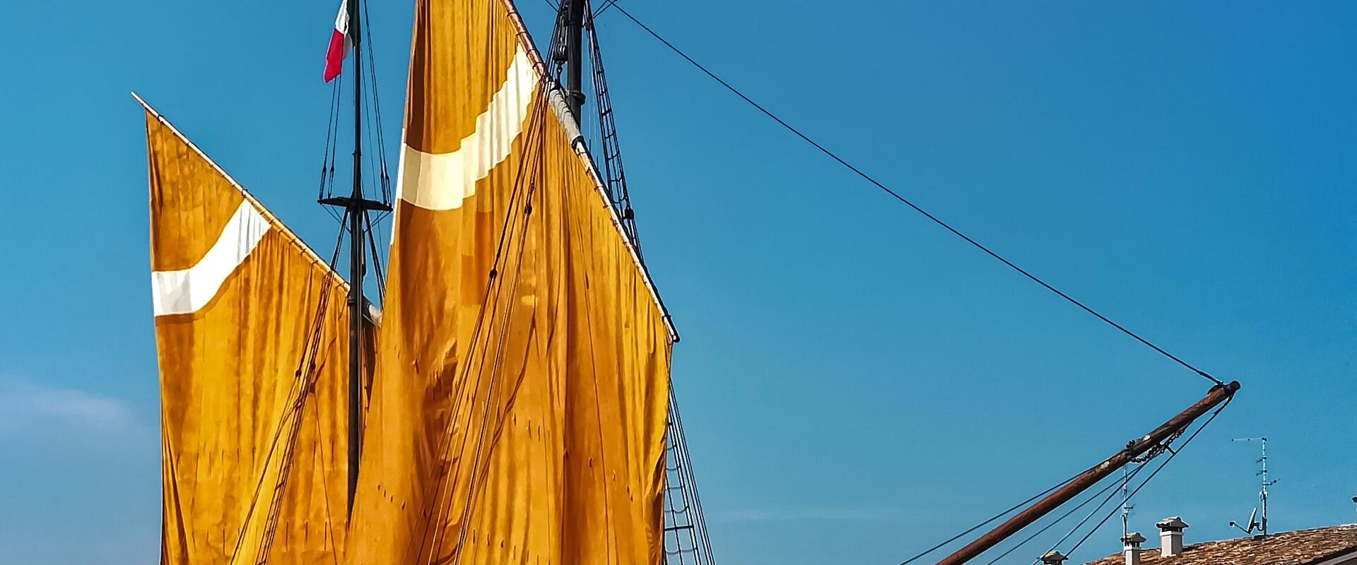 A traditional sailing boat with large yellow sails is moored in a canal harbor, with Italian and EU flags flying, and buildings lining the waterfront under a clear blue sky.