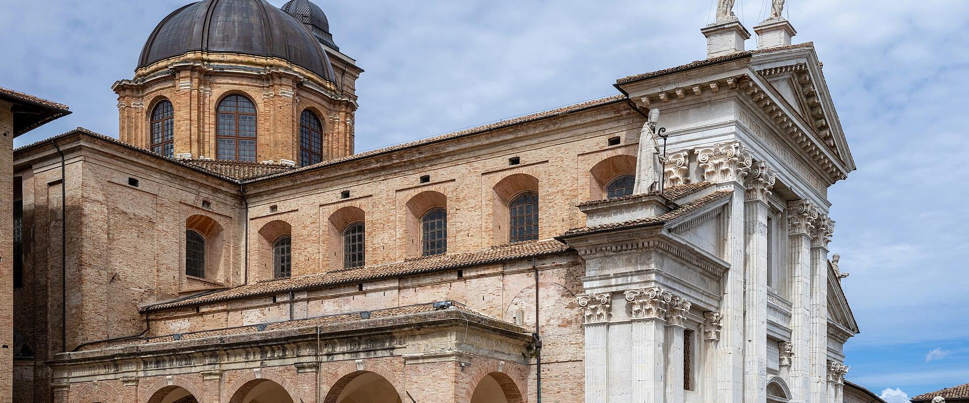 The image shows the exterior of a large historic church with a prominent brick facade, a large central dome, and an ornate white marble portico with columns and statues, set in a paved public square under a partly cloudy sky.