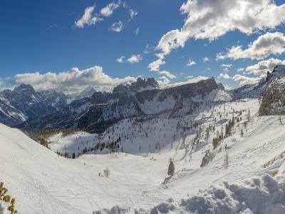 A panoramic winter landscape of the Dolomites featuring the distinctive rock formations of the Cinque Torri (Five Towers) on the left, with snow-covered slopes, scattered larch trees, and distant peaks under a blue sky with scattered clouds.
