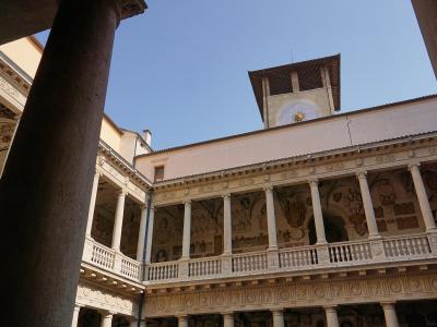 The image shows an interior courtyard of a historic building featuring a two-story loggia with classical columns, ornate friezes, and a prominent clock tower rising above the roofline against a clear blue sky.