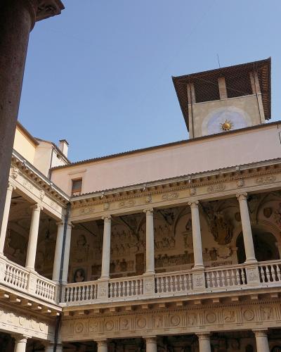 The image shows an interior courtyard of a historic building featuring a two-story loggia with classical columns, ornate friezes, and a prominent clock tower rising above the roofline against a clear blue sky.