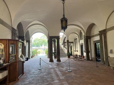 The image shows a covered loggia or courtyard with arched colonnades, terracotta tile flooring, a hanging lantern, display cases on the left, and a view of greenery and stairs through an archway.