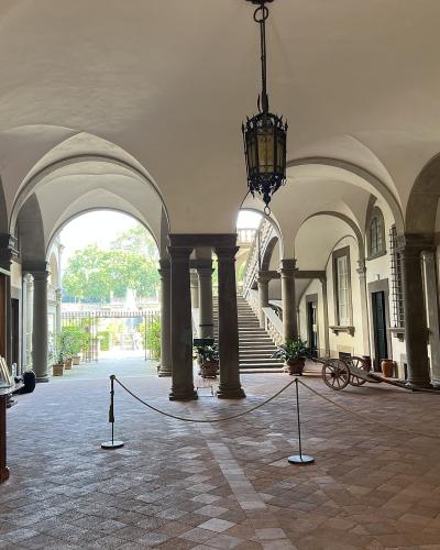 The image shows a covered loggia or courtyard with arched colonnades, terracotta tile flooring, a hanging lantern, display cases on the left, and a view of greenery and stairs through an archway.