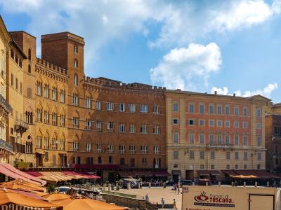 The image shows a sunlit European square with terracotta-colored medieval buildings, including a prominent brick palace with crenellations and arched windows, outdoor café umbrellas in the foreground, and a sign reading "Sapori di Toscana" on a wh