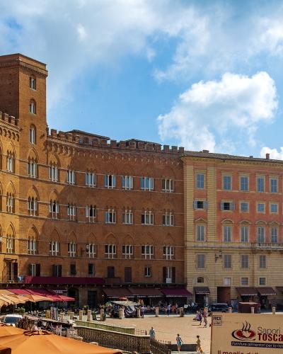 The image shows a sunlit European square with terracotta-colored medieval buildings, including a prominent brick palace with crenellations and arched windows, outdoor café umbrellas in the foreground, and a sign reading "Sapori di Toscana" on a wh