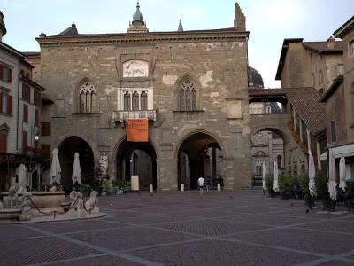 The image shows a large, historic stone building with Gothic-style arched windows and a central balcony, situated in a paved public square; an orange banner reading "Ernesto Neto" hangs from the balcony, and a fountain with lion statues is visible