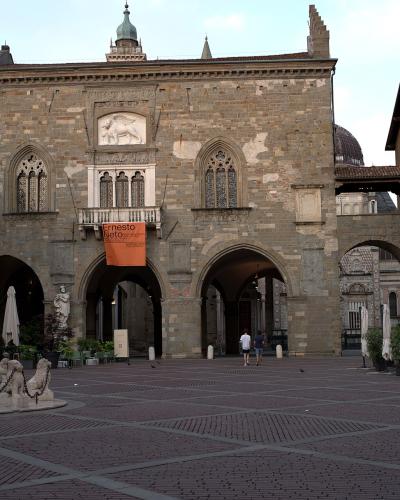The image shows a large, historic stone building with Gothic-style arched windows and a central balcony, situated in a paved public square; an orange banner reading "Ernesto Neto" hangs from the balcony, and a fountain with lion statues is visible