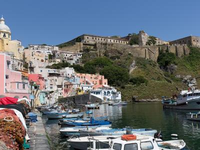 The image shows the colorful harbor of Corricella on the island of Procida, Italy, with fishing boats in the foreground and the fortified medieval village of Terra Murata perched on the hill in the background.