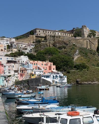 The image shows the colorful harbor of Corricella on the island of Procida, Italy, with fishing boats in the foreground and the fortified medieval village of Terra Murata perched on the hill in the background.