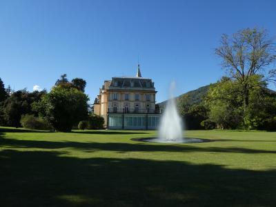 A large, pale-yellow villa with a central tower and mansard roof stands behind a circular fountain on a manicured lawn, framed by trees and a hillside under a clear blue sky.