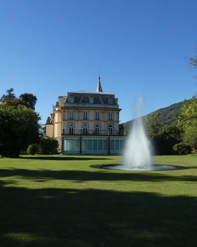 A large, pale-yellow villa with a central tower and mansard roof stands behind a circular fountain on a manicured lawn, framed by trees and a hillside under a clear blue sky.