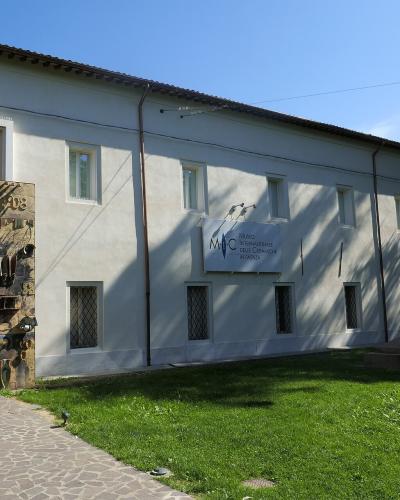 The image shows the exterior facade of a white building with a distinctive stone-clad entrance, a sign reading "MIC MUSEO INTERNAZIONALE DELLE CERAMICHE IN FAENZA", and a bust statue on a pedestal in front of a grassy area.