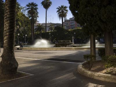 The image shows a large, circular public fountain with multiple water jets spraying upwards, situated in a paved plaza surrounded by tall palm trees, other mature trees, and multi-story buildings in the background under a clear blue sky.