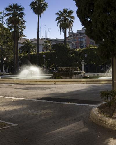 The image shows a large, circular public fountain with multiple water jets spraying upwards, situated in a paved plaza surrounded by tall palm trees, other mature trees, and multi-story buildings in the background under a clear blue sky.