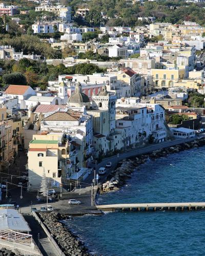 A high-angle view of a densely built coastal town with colorful buildings, a church with a dome and bell tower, a stone breakwater, and a long pier extending into the sea, with hills covered in vegetation and more buildings rising behind.