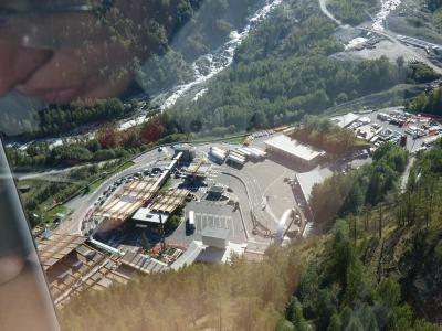 An aerial view looking down at the Mont Blanc Tunnel entrance and toll plaza area in Courmayeur, Italy, with a reflection of a person's face visible in the upper left corner.