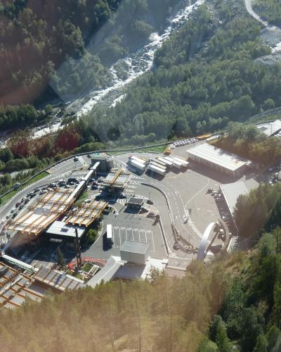 An aerial view looking down at the Mont Blanc Tunnel entrance and toll plaza area in Courmayeur, Italy, with a reflection of a person's face visible in the upper left corner.