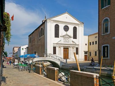 A white Renaissance-style church with a triangular pediment and arched windows stands beside a canal with a small stone bridge, outdoor café seating, and brick buildings under a blue sky.