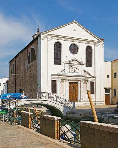 A white Renaissance-style church with a triangular pediment and arched windows stands beside a canal with a small stone bridge, outdoor café seating, and brick buildings under a blue sky.