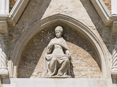 A stone statue of a seated figure in religious vestments, set within a Gothic-style arched niche on an exterior stone wall.