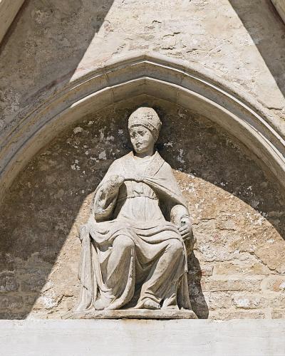 A stone statue of a seated figure in religious vestments, set within a Gothic-style arched niche on an exterior stone wall.