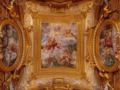 An ornate, gilded ceiling interior featuring Baroque-style frescoes, stucco work, and sculpted figures within elaborate golden frames.