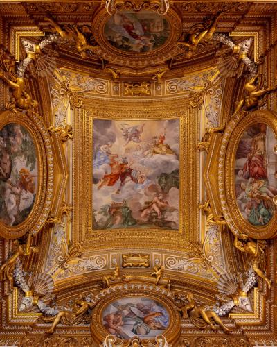 An ornate, gilded ceiling interior featuring Baroque-style frescoes, stucco work, and sculpted figures within elaborate golden frames.
