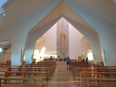 The interior of a modern church with white walls, wooden pews, and a large mosaic depicting the Assumption of Mary at the altar.